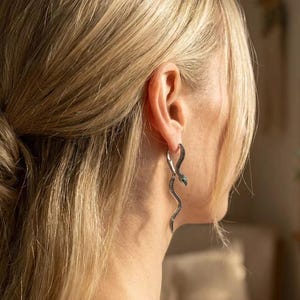 Puede incluir: Un pendiente de serpiente en tono plateado con un cuerpo curvo y alargado, que presenta una superficie texturizada de color oscuro. El diseño de la serpiente cuelga del lóbulo, con la cabeza en la parte superior y la cola curvándose hacia abajo.