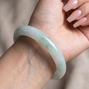 May include: A smooth, polished jade bangle bracelet with a translucent, pale green appearance. The circular accessory features subtle variations in color and a glossy finish, showcasing a solid, continuous band design worn around the wrist.