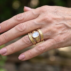 Puede incluir: Un conjunto de tres anillos dorados en un dedo. El anillo central presenta una piedra cabujón blanca iridiscente de forma ovalada. Los dos anillos exteriores tienen una superficie texturizada y grabada, mientras que el anillo central es liso.