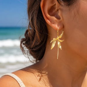 Puede incluir: Un pendiente de tono dorado que presenta un delicado diseño floral de varios pétalos con un tallo largo y delgado. El metal tiene un acabado mate texturizado y la pieza tiene forma de orquídea o lirio estilizado, colgando verticalmente.