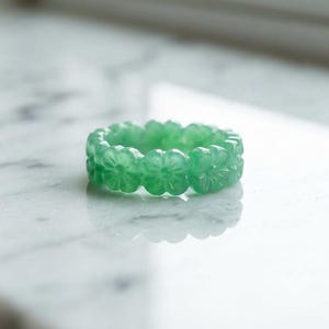 May include: A translucent light green ring carved from a single piece of stone. The band features a continuous pattern of small, five-petaled flowers encircling the entire circumference, creating a textured, floral-inspired design.