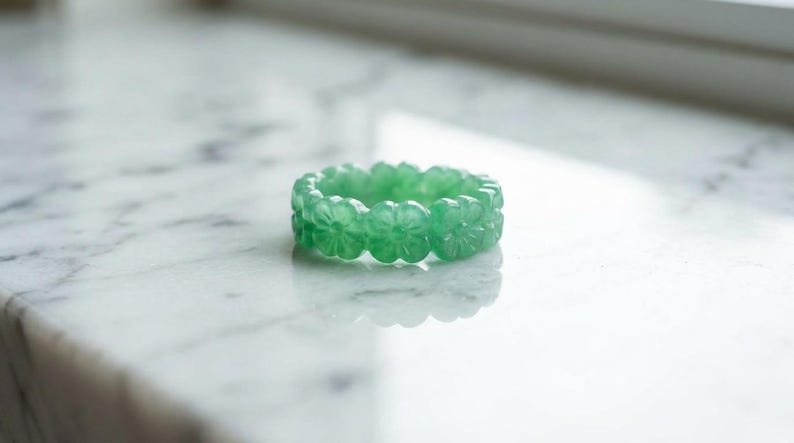 May include: A translucent light green ring carved from a single piece of stone. The band features a continuous pattern of small, five-petaled flowers encircling the entire circumference, creating a textured, floral-inspired design.