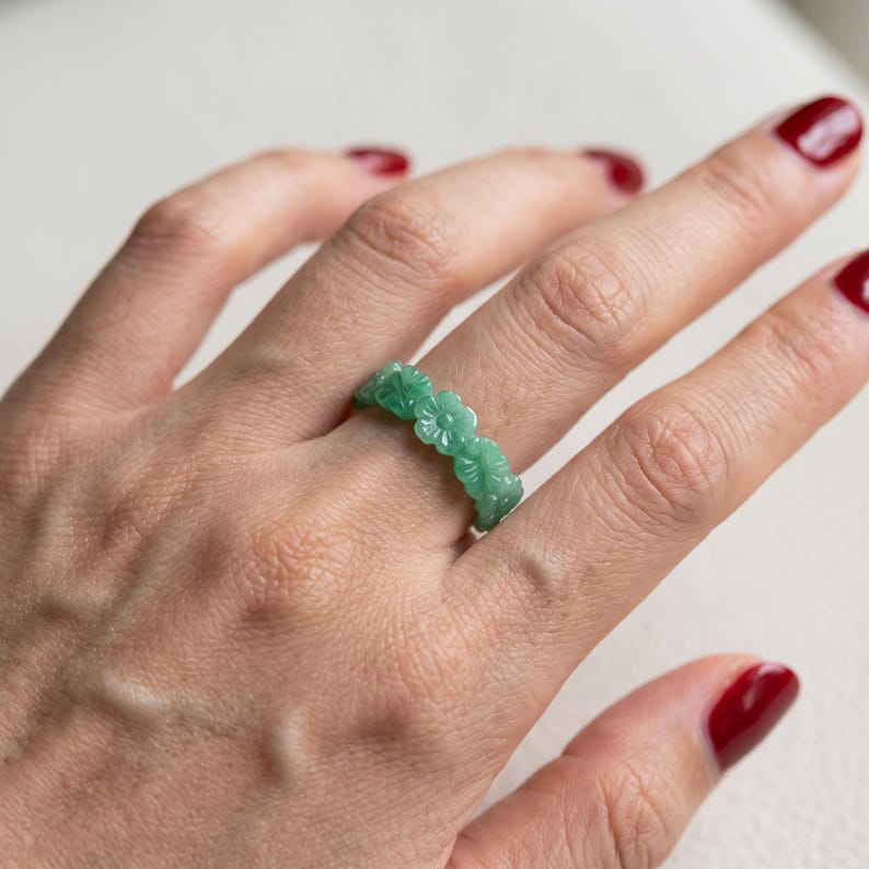 May include: A green ring carved from a single piece of stone, featuring a continuous band of small, detailed flower shapes linked together. The ring has a smooth, polished finish and a translucent quality, showcasing a vibrant, light green color.