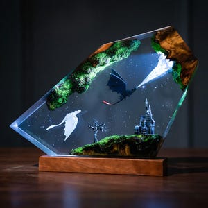 May include: A trapezoidal resin lamp featuring a dark dragon and a white dragon flying above a miniature castle and a bare tree. The scene is encased in clear resin with green moss accents and is mounted on a rectangular wooden base with internal lighting.