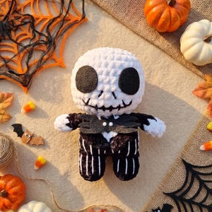 May include: A plush doll featuring a round white head with large black felt eyes and a stitched mouth, wearing a black suit with white vertical stripes and a black felt bat-shaped bowtie. The doll is crafted from soft yarn with a textured finish.