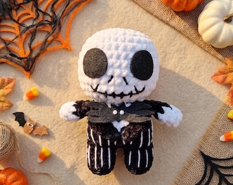May include: A plush doll featuring a round white head with large black felt eyes and a stitched mouth, wearing a black suit with white vertical stripes and a black felt bat-shaped bowtie. The doll is crafted from soft yarn with a textured finish.