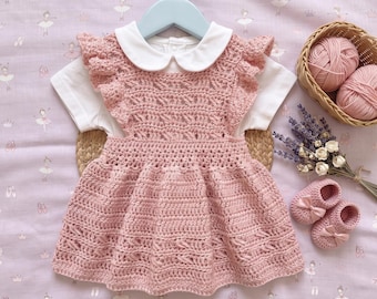 May include: A dusty pink crocheted pinafore dress with ruffled shoulders layered over a white short-sleeved shirt with a rounded collar. The set includes a pair of matching pink crocheted booties with small bows, displayed with two balls of yarn.