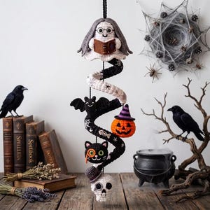 May include: A vertical crochet hanging decoration featuring a ghost with glasses holding a book, a black bat, an orange jack-o'-lantern with a purple hat, a black cat with an orange monocle, and a white skull with a top hat and a monocle.