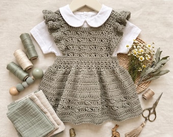 May include: A sage green crocheted pinafore dress layered over a white short-sleeved shirt with a rounded collar. The dress features ruffled shoulder straps and a textured stitch pattern. Accessories include spools of thread, fabric, and scissors.