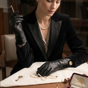 Peut inclure: Gants en cuir noir avec boutons dorés sur les poignets, portés avec un blazer en velours noir, une chemise à col crème et un collier de perles. Les gants sont présentés avec un collier de perles, deux clés en laiton et un écrin en velours.