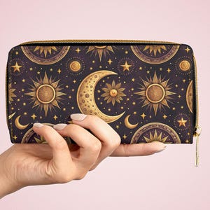 Peut inclure: Un portefeuille rectangulaire à fermeture éclair avec un fond sombre orné d'un motif répétitif de soleils, de croissants de lune et d'étoiles dorés. Le portefeuille est doté d'une fermeture éclair en métal doré sur le dessus et le côté.