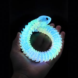 May include: A glowing, articulated snake-like toy with a segmented body that emits a bright blue and light green light. The flexible, multi-jointed creature features a smooth, rounded head with a single visible eye opening, coiled into a circle.