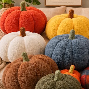 Puede incluir: Una colección de cojines con forma de calabaza hechos de tela sherpa texturizada. Las calabazas vienen en varios colores, incluyendo naranja, amarillo, blanco, azul, marrón y verde oliva, cada una con un pequeño tallo en la parte superior.