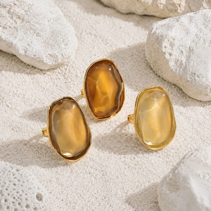 Puede incluir: Tres anillos con piedras de resina translúcida de color ámbar, de formas irregulares y lisas, montadas en bandas de metal dorado. Cada anillo presenta una forma orgánica única con tonos variables de amarillo y marrón.