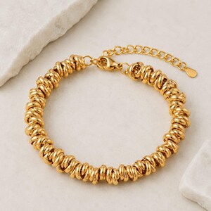 Puede incluir: Una pulsera de tono dorado compuesta por una serie continua de pequeños eslabones circulares interconectados. La pulsera cuenta con un cierre de mosquetón y una cadena de extensión ajustable que termina en un pequeño dije metálico en forma de gota.