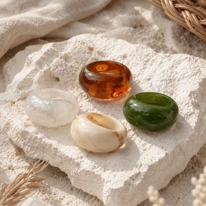 Puede incluir: Cuatro anillos gruesos con un diseño redondeado y liso. Un anillo es de color ámbar translúcido, otro es verde oscuro sólido, otro es de color crema veteado y otro es blanco nublado semitranslúcido. Cada anillo tiene una banda gruesa y pulida.