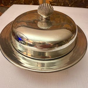 May include: A silver-toned metal butter dish with a circular base and a matching domed lid. The lid features a decorative handle shaped like a seashell. The polished surface of the metal reflects the surrounding environment and light.