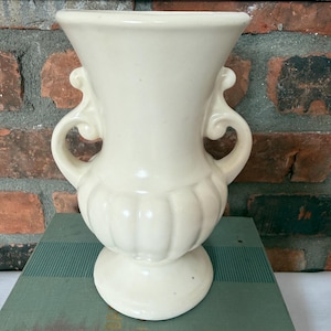 May include: A cream-colored ceramic vase featuring a flared rim, a bulbous body with vertical fluted detailing, and two symmetrical scroll-shaped handles on the sides. The vase stands on a circular pedestal base and has a smooth, glossy finish.