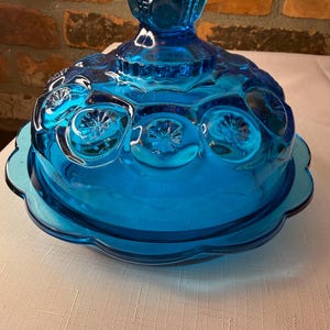 May include: A vibrant blue glass butter dish featuring a domed lid with a moon and star pattern and a scalloped edge base. The lid is topped with a decorative knob handle, and the entire piece is crafted from translucent, deep blue pressed glass.
