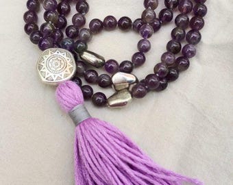 May include: A beaded necklace featuring polished dark purple amethyst spheres, interspersed with three smooth metallic silver-toned accents. A circular silver-toned charm with an etched sunburst pattern connects to a long, vibrant lavender tassel.