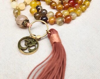 May include: A mala necklace featuring multi-colored, semi-translucent stone beads in shades of yellow, orange, brown, and cream. It includes a gold-toned metal Om charm, a circular connector, and a long, dusty rose-colored fabric tassel.