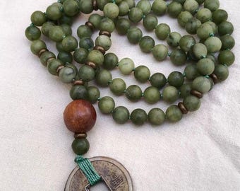 May include: A mala necklace featuring numerous round, mottled olive-green stone beads strung on green cord. The necklace includes small metallic spacers, a single large polished wooden bead, and a circular metal coin pendant with text.