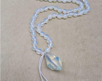 May include: A long necklace featuring translucent, milky white opal-like beads strung on a white cord. The necklace ends in a faceted, teardrop-shaped pendant and two smaller dangling bead accents. A small logo with the text SACRED is visible.