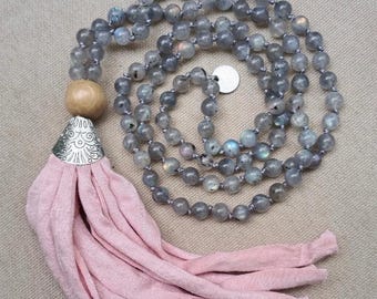 May include: A beaded necklace featuring round grey labradorite beads, a single smooth wooden bead, and a silver-toned metal cap with engraved floral patterns. A long, soft pink fabric tassel hangs from the cap. The text SACRED is visible.