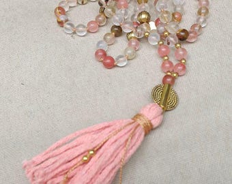 May include: A beaded necklace featuring translucent pink, clear, and golden-brown stone beads interspersed with small gold-toned metal spacers. A large, textured gold-toned metal bead connects to a long, soft pink cotton tassel.