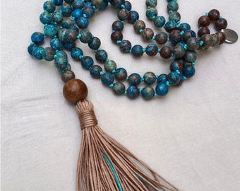 May include: A beaded necklace featuring round blue and brown marbled stone beads strung on turquoise cord. A large, smooth, dark brown wooden bead sits above a long, beige-colored tassel. A small silver-toned circular charm is attached at the end.
