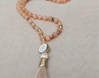 May include: A long necklace featuring peach-colored round beads interspersed with small metallic silver spacers. The necklace includes a circular white and gold charm and a long, light beige fabric tassel at the bottom end.