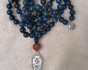 May include: A necklace featuring dark blue, spherical beads arranged in a circular pattern. A single brown wooden bead sits above a silver-toned, shield-shaped metal pendant with intricate cut-out designs. A small, round silver charm is attached.