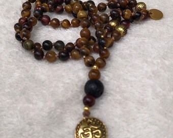 May include: A beaded necklace featuring brown and black tiger eye beads, a single textured black lava bead, and gold-toned metal accents. The necklace includes a round gold-toned pendant embossed with the Om symbol and a small circular charm.
