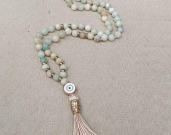 May include: A long beaded necklace featuring pale green and cream-colored round beads interspersed with small gold-toned spacers. The necklace includes a circular charm with an eye motif and a long, light beige fabric tassel at the end.