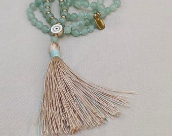 May include: A mala necklace featuring light green gemstone beads, small gold-toned spacer beads, and a central white circular charm with a black eye symbol. A long, beige-colored thread tassel hangs from the bottom, secured by light blue thread.