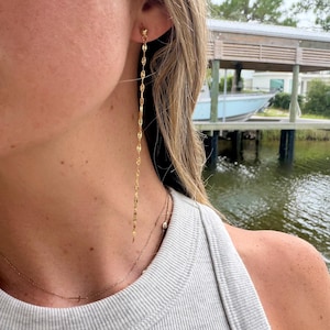 May include: A long, gold-toned chain earring featuring a delicate, elongated link design that hangs vertically from the earlobe. The earring is crafted with a simple stud post backing and displays a polished, reflective metallic finish throughout.