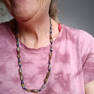 May include: A long necklace featuring a repeating pattern of smooth brown oval beads, dark blue cylindrical beads with thin yellow and white stripes, and small dark blue round beads strung on a dark cord, resting against a pink tie-dye shirt.