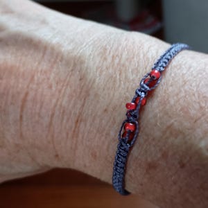 May include: A grey cord bracelet featuring a series of small, vibrant red beads integrated into the woven structure. The bracelet is constructed with a knotted design, showcasing a repeating pattern of grey thread loops that secure the red beads.