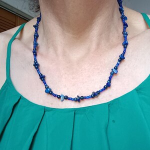 May include: A necklace featuring a series of small, deep blue, irregular stone chips interspersed with tiny, round, dark blue glass beads. The beads are strung together in a repeating pattern, creating a textured, multi-faceted appearance around the neck.