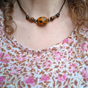 May include: A necklace featuring a dark brown leather cord with a central oval orange bead decorated with black spots, flanked by two smaller brown beads and two metallic ribbed spacers, secured with simple knots on each side of the focal beads.