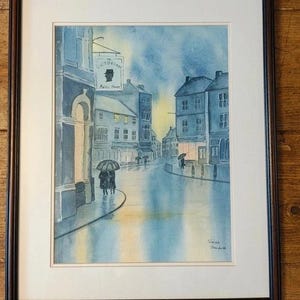 May include: A framed watercolour painting depicting a rainy street scene at dusk with blue and yellow tones. The artwork features buildings, figures with umbrellas, and a sign that reads The Victorian Relic House. The painting is signed Sarah Stanforth.