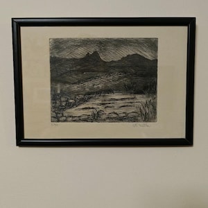 May include: A framed black and white etching print depicting a landscape with a body of water in the foreground, rolling hills, and a mountain range under a dark, hatched sky. The print is numbered 2/50 and signed by the artist in pencil.