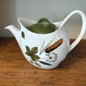 Op de afbeelding: Een witte keramische theepot met een gebogen handvat en tuit, voorzien van een botanische print van groene bladeren, bruine lisdodden en een paardenbloempluis. De theepot heeft een afneembaar olijfgroen deksel met een kleine knop.