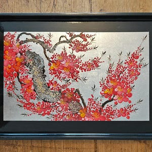 May include: A rectangular black serving tray featuring a central decorative panel with a silver background. The panel displays a painted illustration of a flowering tree branch with vibrant red and orange blossoms and dark textured bark.