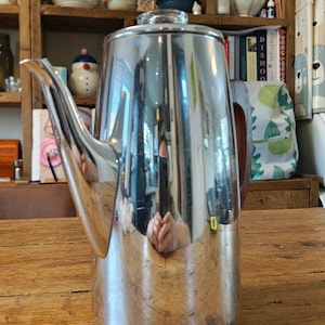 May include: A tall, cylindrical coffee pot made of polished, reflective stainless steel. It features a long, curved spout on the left side, a dark brown wooden handle on the right, and a clear glass knob on the lid. The surface reflects the surroundings.