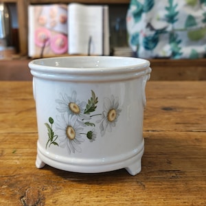 May include: A white ceramic planter with a cylindrical shape, featuring a delicate floral pattern of white daisies with yellow centres and green leaves. The planter stands on three small feet and has a smooth, glossy finish with subtle crackle details.