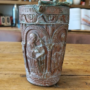 May include: A cylindrical vessel with a weathered, rust-coloured finish and teal patina accents. The exterior features intricate relief carvings of figures in arched panels, Latin text inscriptions, and decorative borders around the top and base.