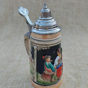 May include: A ceramic beer stein featuring a metallic lid with a thumb lever. The body is black with a colourful, raised relief scene depicting two figures in traditional attire. The base and rim are a light tan colour, and the handle is curved.