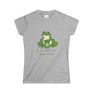 Könnte beinhalten: Ein graumeliertes Kurzarm-T-Shirt mit einer grünen Frosch-Illustration, der einen grünen Recycling-Behälter hält. Unter dem Frosch steht der Text "IT'S NOT THAT EASY BEING GREEN" in einer dunkelbraunen, handgezeichneten Schrift mittig.