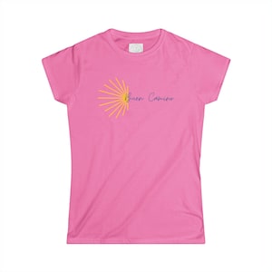 May include: A bright pink short-sleeved t-shirt featuring a yellow sunburst graphic on the left side. The text "Buen Camino" is printed in a dark blue cursive script next to the sunburst design. The shirt has a crew neckline and a standard fit.
